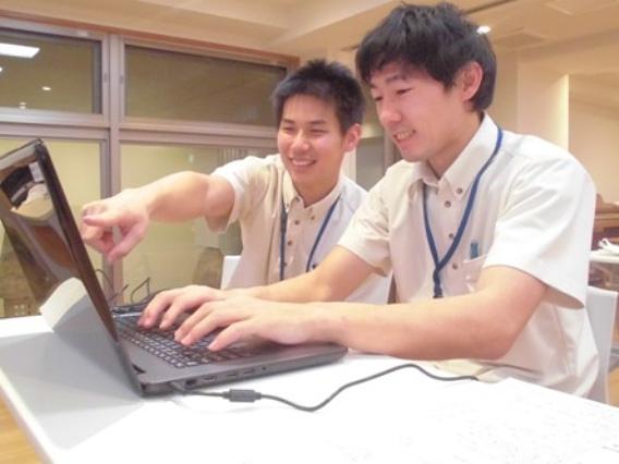 写真手前。新人スタッフが先輩スタッフから社内システムについて教わっています。これから頑張ります！